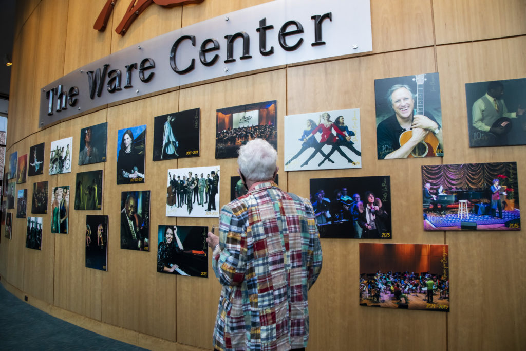 A white-haired visitor walks through a gallery wall depicting previous shows at the Ware Center